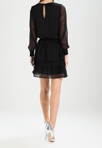 Vrouw in een zwart doorzichtig jurk met polka-dots, lange mouwen en een gelaagde rok, gezien vanaf de achterkant, met zilveren hoge hakken.