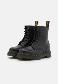 Stivali alla caviglia in pelle nera con punta rotonda, design con lacci, suola in gomma testurizzata, cuciture gialle e una linguetta con logo sul retro.