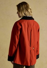 Czerwona kurtka o prostokątnym kształcie, z czarnym kołnierzem, guzikami jako ozdobami oraz rozcięciem z tyłu. Materiał wydaje się gładki i matowy.