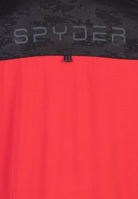Chaqueta deportiva roja con un patrón de camuflaje negro en la parte superior. Presenta la marca "SPYDER" en letras grises. Textura suave, diseño moderno.