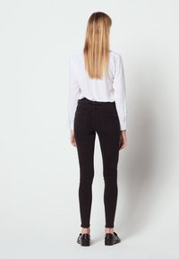 Jeans negros ajustados de talle alto, con bolsillos traseros y un corte slim. Combinados con una camisa blanca de manga larga y zapatos negros.