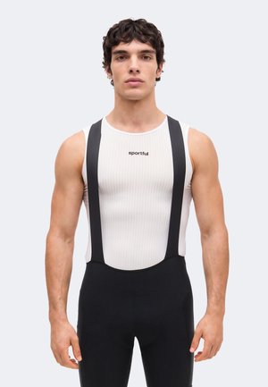 Atlétikus férfi fehér, ujjatlan bordás felsőt visel „sportful” felirattal és fekete kerékpáros nadrágot vállpántokkal, egyszínű háttér előtt.