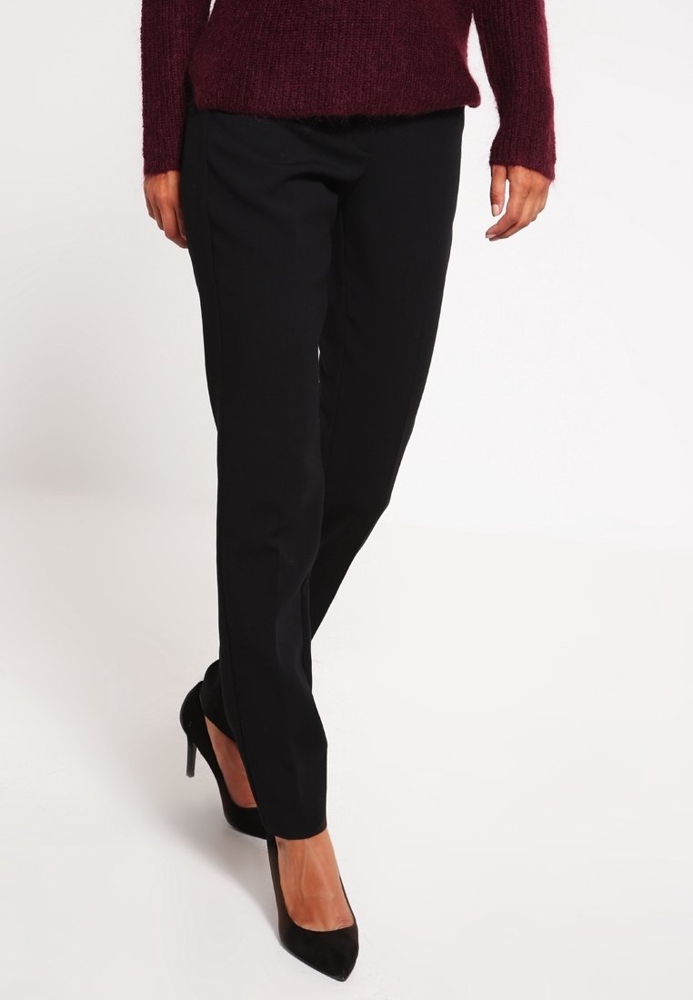 Femme portant un pantalon noir slim, des talons hauts noirs, et un pull en maille bordeaux, debout devant un fond blanc.