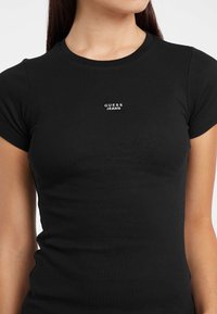 T-shirt nera a maniche corte con design aderente. Presenta il logo "GUESS JEANS" in bianco nella zona del petto. Tessuto morbido.