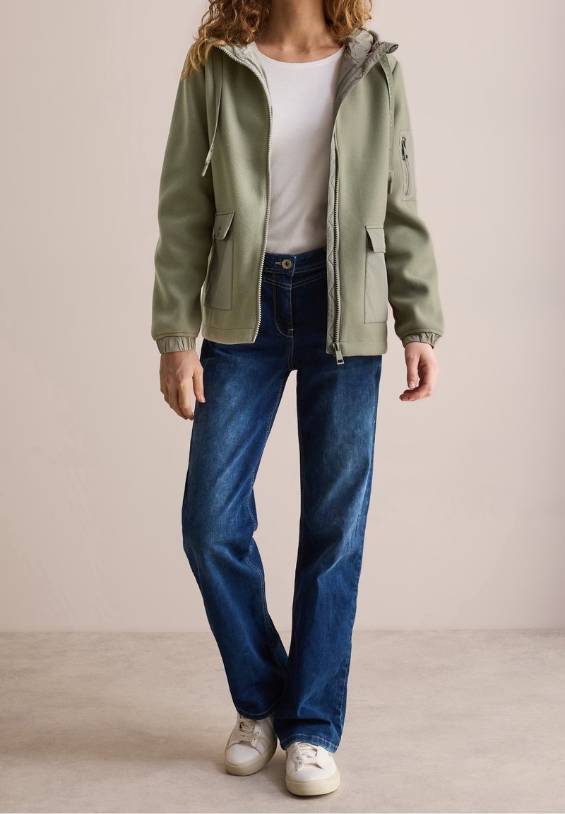 Femme portant une veste zippée verte, un t-shirt blanc, un jean bleu et des baskets blanches, debout sur un fond uni.