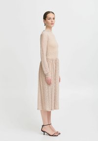 Langärmliges, knielanges Kleid aus hellem, beige strukturiertem Spitzenstoff mit tailliertem Sumpf und ausgestellt Rock, kombiniert mit schwarzen, knöchelhohen Sandalen.