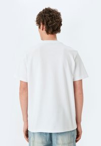 Hvid kortærmet t-shirt med rund hals, der fremviser et struktureret stof. Bagsiden har en glat finish og et afslappet design. Lys baggrund.