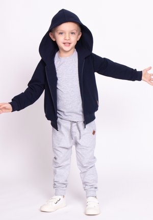 Granatowa bluza z kapturem na zamek błyskawiczny, z podszytym kapturem, szara koszulka i szare spodnie dresowe. Strój uzupełniają białe sneakersy z ciemnymi paskami i dodatkami.