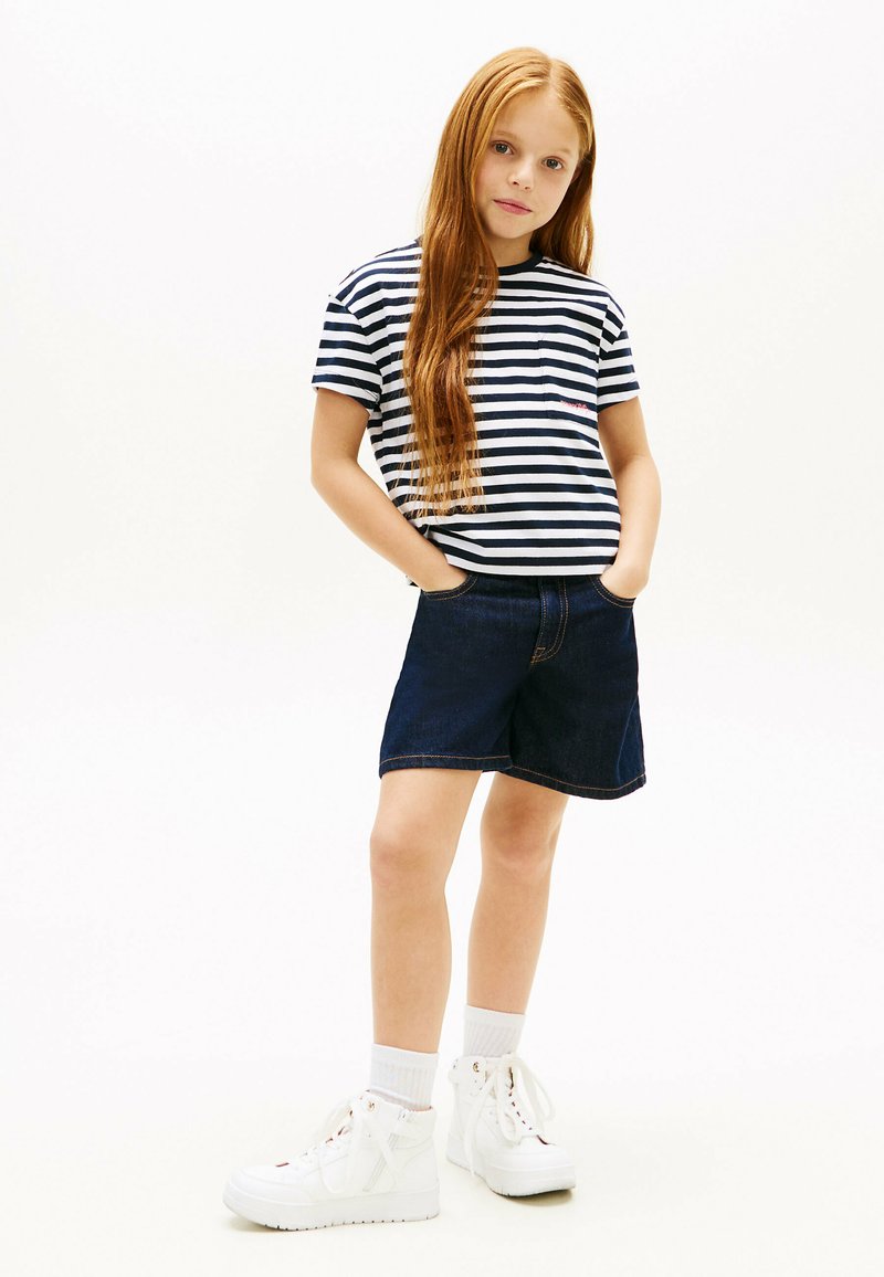 Jeune fille aux longs cheveux roux portant un t-shirt à rayures bleu marine et blanc, un short en jean foncé, des chaussettes blanches et des baskets montantes blanches.
