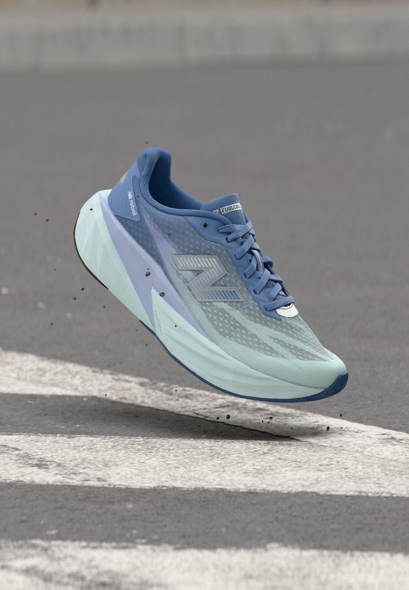 Chaussure de course New Balance bleue et bleu clair en suspension au-dessus d’un bitume avec marquages blancs et petites particules de saleté.
