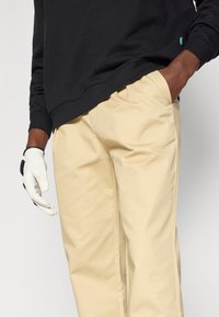 Melnas jakas ar noapaļotu kakla izgriezumu, bēšas chinos bikses ar gludu faktūru un baltas cimdi ar melniem akcentiem, kas turēti relaksētā pozā.