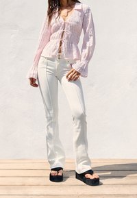 Light pink, textured blouse with a V-neck and bell sleeves, paired with white flared jeans and black platform sandals.