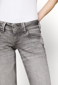 Donna che indossa jeans grigi sbiaditi con chiusura a doppio bottone e una canotta bianca aderente, con la mano appoggiata sull'anca su uno sfondo bianco.