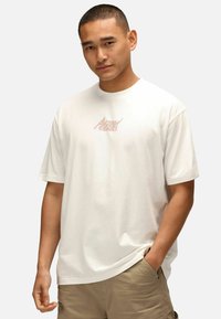 Weißes T-Shirt mit kurzen Ärmeln aus weichem Baumwollstoff, das auf der Brust ein grafisches Motiv mit "Action Comics" in Pastellfarben zeigt.