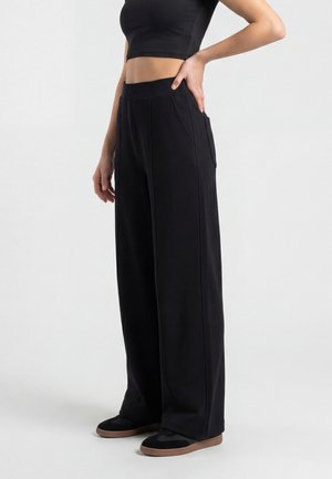 Persona che indossa pantaloni neri a gamba larga e top corto nero, in piedi con una mano sui fianchi contro uno sfondo semplice.