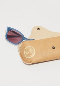 Gafas de sol azul con forma de ojo de gato y lentes degradados en color púrpura, dentro de un estuche de cuero marrón claro con un logo en relieve dorado y cierre a presión.