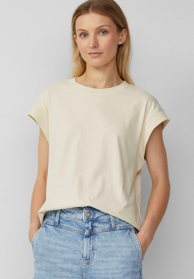 Crème-kleurig katoenen T-shirt met opgerolde korte mouwen, ronde halslijn en een relaxte pasvorm, gecombineerd met lichtblauwe spijkerbroek.