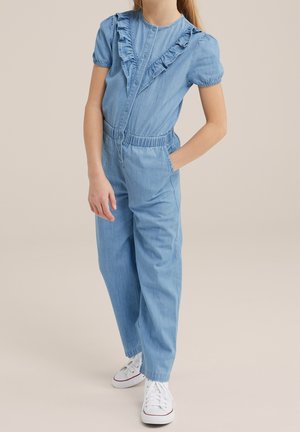 Combinaison en denim bleu clair à manches courtes, devant à volants, taille élastique et poches latérales. Associée à des baskets blanches.