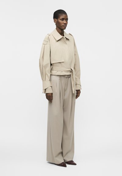 Γυναίκα που φοράει μπεζ cropped σακάκι με ζώνη και φαρδιά μανίκια και μπεζ ψηλόμεσα φαρδιά παντελόνια, σε συνδυασμό με σκούρα μυτερά παπούτσια.