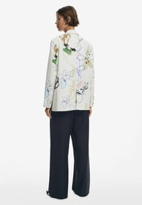 Blazer imprimé floral blanc avec des designs abstraits, présentant des manches longues et une coupe ajustée, associé à un pantalon large noir.