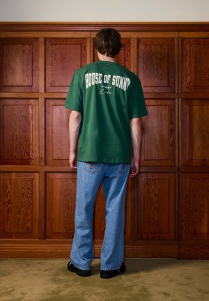 Egy személy áll, háttal egy fa borítású falnak, zöld "House of Sunny" pólót, kék farmert és fekete cipőt visel a szőnyeggel borított padlón.