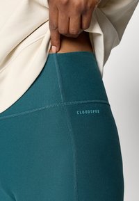 Leggings teal com uma textura suave, apresentando costuras costuradas e um logo na lateral. Usados sob uma blusa de cor clara.
