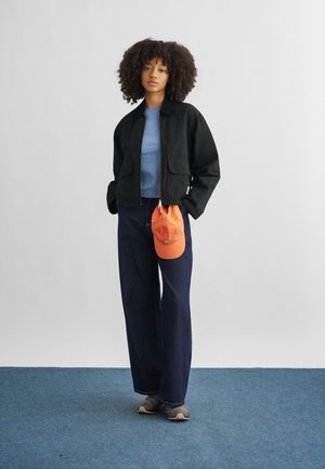 Kvinde med krøllet hår iført sort jakke, blå skjorte, jeans med brede ben, grå sko, holder en orange kasket, står på blå tæppe foran hvid væg.