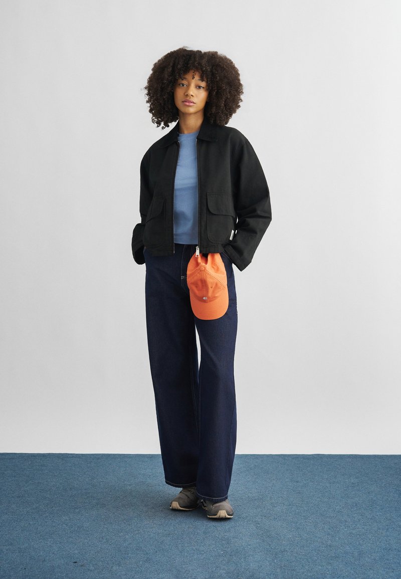 Woman with curly hair wearing black jacket, blue shirt, wide-leg jeans, gray shoes, holding an orange cap, standing on blue carpet against white wall.