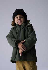 Giacca isolante verde con cerniera nera, cappuccio foderato in pelliccia e polsini elasticizzati, abbinata a pantaloni cargo marroni e un berretto di maglia nero.