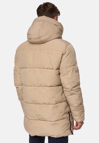 Beige pufferjakke med hette, med horisontal quiltet mønster, en avslappet passform og glidelåslommer på siden. Materialet virker glatt og lett.