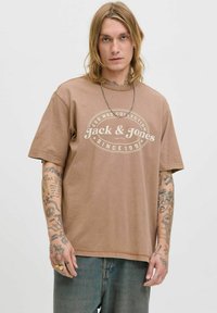 Brun bomulds t-shirt med korte ærmer, prydet med et hvidt cirkulært logo-print. Sammen med lyseblå denimjeans. Casual pasform.