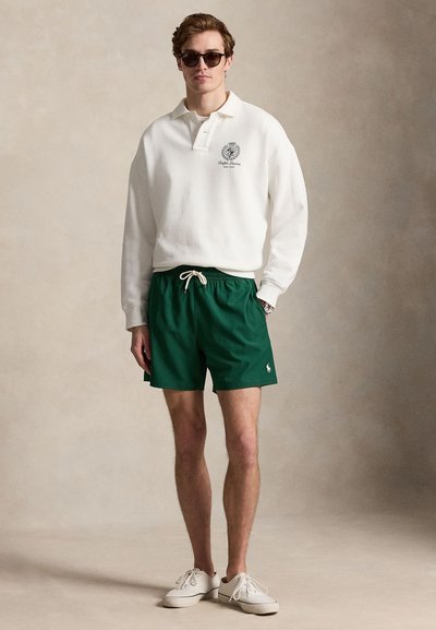 Homme portant un polo blanc à manches longues, un short vert à cordon, des baskets blanches et des lunettes de soleil, debout devant un fond beige uni.