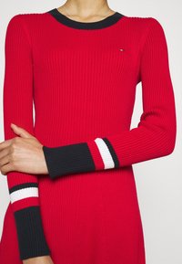 Punainen ribattuja mekko, jossa on pyöreä musta kaulus ja pitkät hihat. Hihoissa on mustat, valkoiset ja punaiset raidalliset yksityiskohdat.