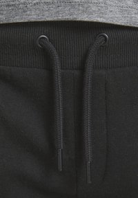 Schwarze Sweatpants mit einem gerippten Bund, verstellbaren Kordeln und einer strukturierten Oberfläche, teilweise bedeckt durch den Saum eines grauen Shirts.