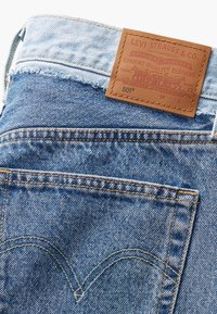 Jean Levi's en denim bleu montrant les coutures de la poche arrière et un patch en cuir marron avec logo et texte sur la ceinture.
