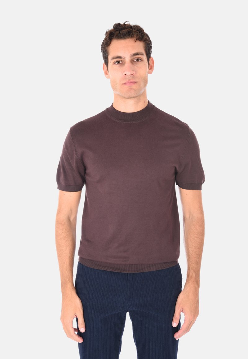 Uomo con capelli ricci corti che indossa un maglione corto a maniche corte di color marrone e pantaloni a coste blu scuro, in piedi davanti a uno sfondo chiaro e uniforme.