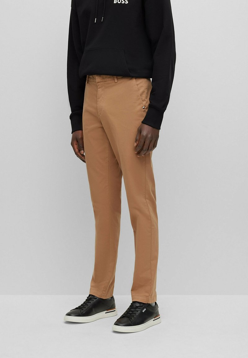 Homme portant un pantalon slim beige, un sweat à capuche noir et des baskets noires à semelles blanches, debout devant un fond gris uni.