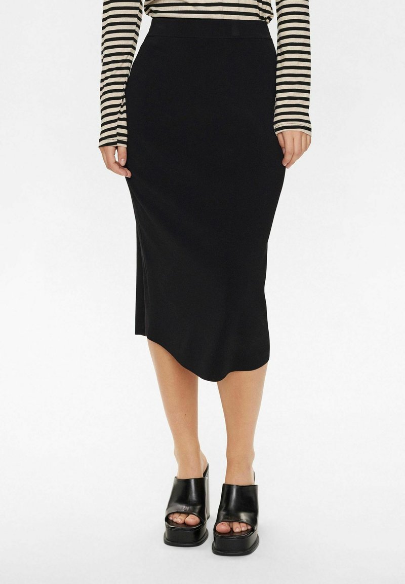 Femme portant une jupe midi noire, des chaussures plateforme noires à bout ouvert et un top à manches longues beige et noir rayé sur fond blanc.