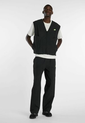 Mann steht mit verschränkten Händen hinter dem Rücken und trägt eine schwarze Weste mit Reißverschluss über einem weißen T-Shirt, schwarze Hose und schwarze Sneakers vor weißem Hintergrund.