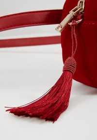 Czerwona zamszowa torebka z złotym zamkiem błyskawicznym, ozdobiona plecionym chwostem i eleganckim czerwonym paskiem.