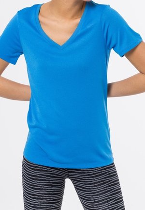 Person iført en knaldblå kortærmet V-hals skjorte og sorte og grå stribede leggings, med hænderne hvilende på hofterne.