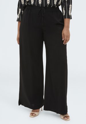 Donna che indossa pantaloni neri a gamba larga con vita elasticizzata e coulisse, abbinati a tacchi beige open toe e una maglia con fantasia.