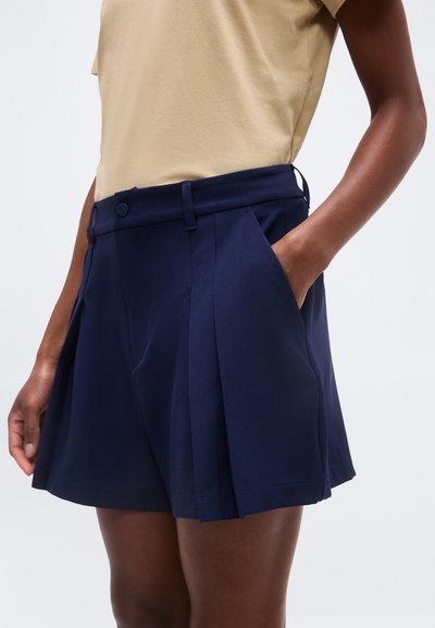 Άτομο που φοράει ναυτικό σορτς με πιέτες και τσέπες, συνδυασμένο με μπεζ κοντομάνικο τοπ, στέκεται μπροστά σε ένα απλό φόντο.