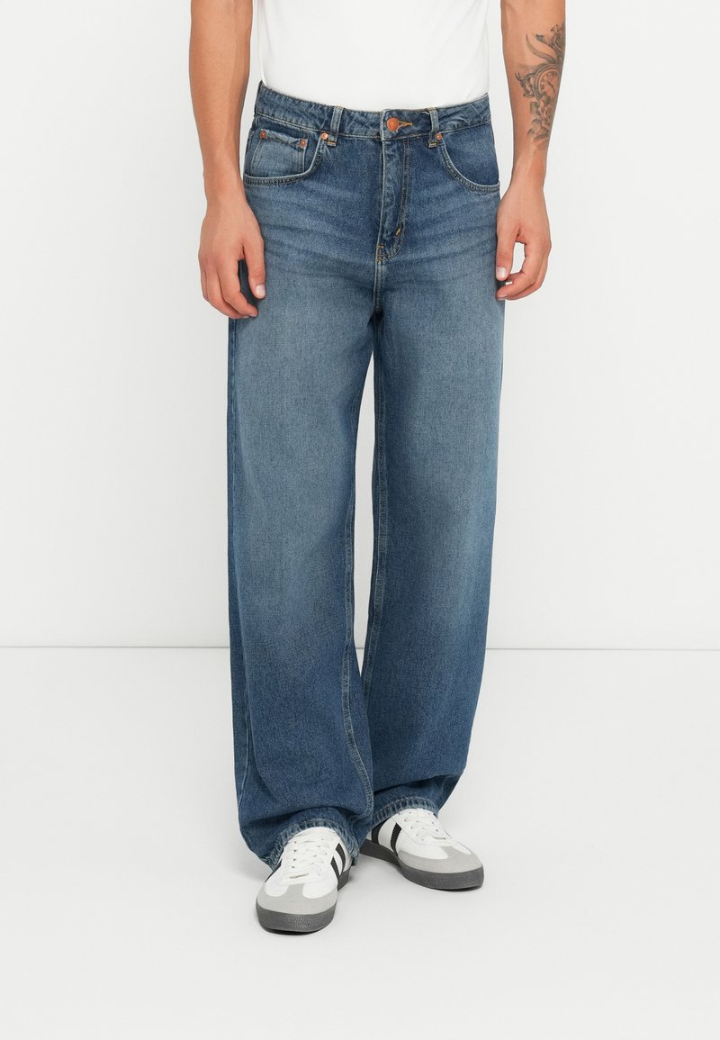 Lyseblå vide jeans laget av denim, med fem-lommedesign, knappelukk og detaljer i oransjefarge. Kombinert med hvite og grå joggesko.