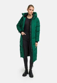 Lungo cappotto imbottito verde con cappuccio, dotato di chiusura a zip, tasche laterali e polsini a costine. Indossato sopra una maglia nera e jeans scuri.