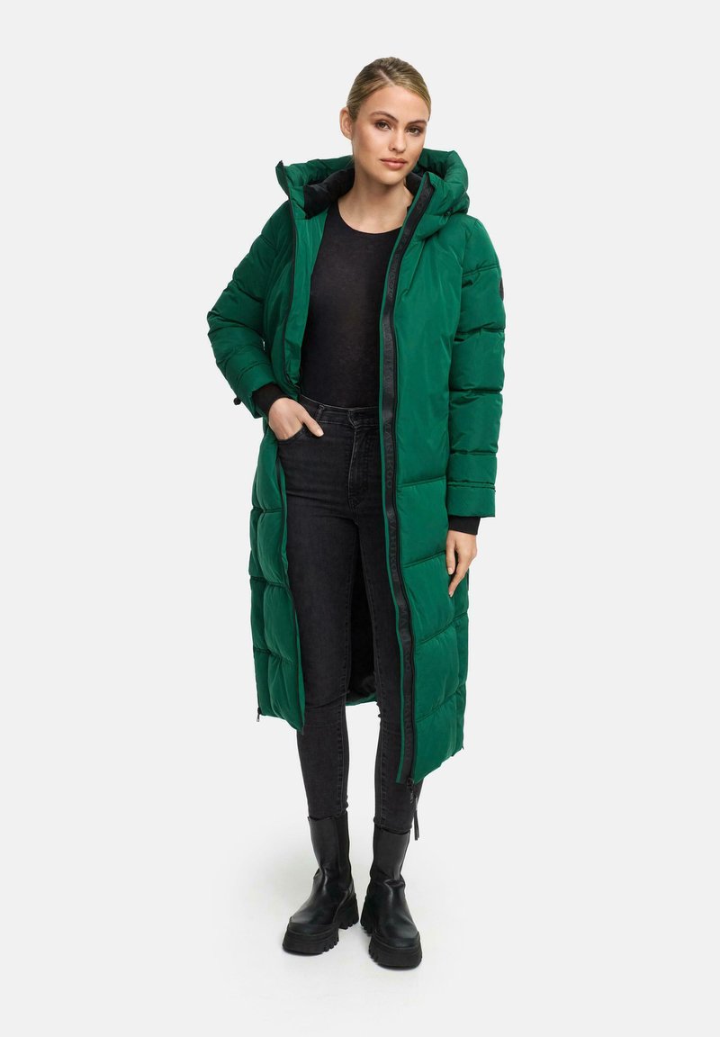 Abrigo acolchado largo verde con capucha, con cierre de cremallera, bolsillos laterales y puños acanalados. Lleva puesto encima de una camiseta negra y unos jeans oscuros.