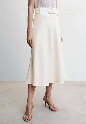Femme portant une jupe midi crème avec une large ceinture et une boucle ronde, associée à des sandales à talons hauts beiges à lanières et un top sans manches côtelé.