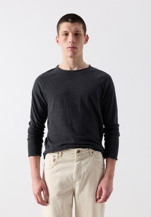 Noor mees seisab näoga otse, kandes tumedat pika varrukaga särki ja heledaid pükse, taustaks on ühtlane valge taust.