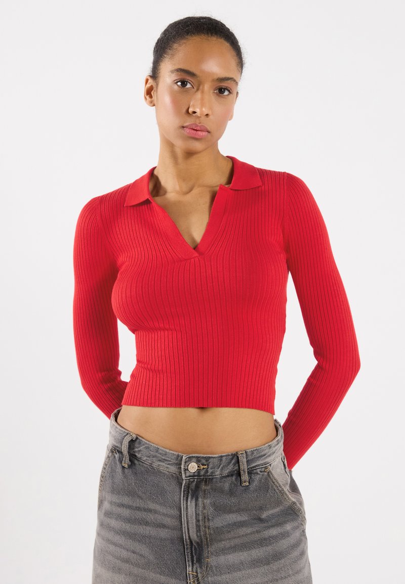 Rotes, geripptes Strickoberteil mit langen Ärmeln, V-Ausschnitt und Kragen, kombiniert mit hoch geschnittenen, verwaschenen Jeans, die eine glatte Textur und eine figurbetonte Passform zeigen.