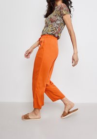 Pantalons courts orange, légers, avec une taille élastique et des poches latérales. Associés à un haut à manches courtes à motifs et à des sandales.